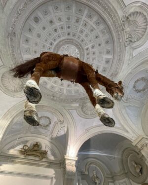 Daniela Collu Instagram - C’è un cavallo imbalsamato appeso in mezzo alla stanza di un museo: è un’opera di Maurizio Cattelan e si intitola Novecento. Ne ho visti un milione di cavalli nei musei, ritratti in posizione rampante, solenni nelle sculture equestri, dipinti nei paesaggi ottocenteschi, morenti nelle scene di guerra di tutte le epoche, a terra, galoppanti, in mandrie, solitari, ma così mai. 
Il collo è deformato, allungato dalla gravità e dalla tassidermia, le zampe a guardarle da sotto sembrano più tozze, più lunghe e larghe, e un senso di precarietà pervade i sensi, mentre si attraversa la stanza sotto questo peso galleggiante, immobile, inerme. Si chiama come il secolo in cui siamo nati, quello in cui la maggior parte delle sciagure che ora non sappiamo manovrare sono state costruite, un secolo di errori, di scoperte, di sconfitte e vittorie, di grandi passi per l’umanità e di crisi esistenziali senza ritorno. È la natura morta più disorientante, straniante e cruda che si possa immaginare, parla di simboli decaduti, di corpi infranti, di certezze mancate, di fallimenti annunciati. Di quel senso di impotenza e di inerzia umana che ancora, in un nuovo secolo, ci soffoca terribilmente. #unminutodarte