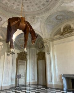 Daniela Collu Instagram - C’è un cavallo imbalsamato appeso in mezzo alla stanza di un museo: è un’opera di Maurizio Cattelan e si intitola Novecento. Ne ho visti un milione di cavalli nei musei, ritratti in posizione rampante, solenni nelle sculture equestri, dipinti nei paesaggi ottocenteschi, morenti nelle scene di guerra di tutte le epoche, a terra, galoppanti, in mandrie, solitari, ma così mai. Il collo è deformato, allungato dalla gravità e dalla tassidermia, le zampe a guardarle da sotto sembrano più tozze, più lunghe e larghe, e un senso di precarietà pervade i sensi, mentre si attraversa la stanza sotto questo peso galleggiante, immobile, inerme. Si chiama come il secolo in cui siamo nati, quello in cui la maggior parte delle sciagure che ora non sappiamo manovrare sono state costruite, un secolo di errori, di scoperte, di sconfitte e vittorie, di grandi passi per l’umanità e di crisi esistenziali senza ritorno. È la natura morta più disorientante, straniante e cruda che si possa immaginare, parla di simboli decaduti, di corpi infranti, di certezze mancate, di fallimenti annunciati. Di quel senso di impotenza e di inerzia umana che ancora, in un nuovo secolo, ci soffoca terribilmente. #unminutodarte Castello di Rivoli Museo d'Arte Contemporanea