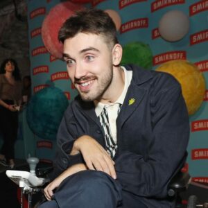 George Robinson Instagram - #ad I had a great time celebrating the launch of @smirnoffeurope WE DO US Live. While I love a good party, it’s way better when everyone can be a part of it. That’s why making nightlife more accessible is so important. @smirnoffeurope and Disability-led organization @tiltingthelens are working together to improve accessibility by supporting the Disabled community in clubs, bars and pubs - more to come! To everyone involved - thank you for such a fabulous night and showing easily it can be done.

************

Image Description: George Robinson is a white, masc wheelchair user. He has green eyes, cropped, dark hair, a moustache and dark stubble along his jawline. He is wearing a dark jacket and chinos and a white and black houndstooth polo shirt. He is wearing thick, textured brown socks and black leather shoes. On his lapel he has a small light green pin. He is positioned in front of a sky blue branding board, with the red and white Smirnoff logo repeated all over and red, green and yellow spheres covered in felt as a decoration. He is smiling for the picture and is at the Smirnoff ‘We Do Us Live’ event, celebrating accessibility in music. The event took place on 29/11 at 26 Leake Street in London.
