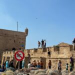 Vera Kolodzig Instagram - Dia 13: Conhecida como a “Cidade Ventosa de África”, Essaouira atrai muitos kitesurfers durante o ano inteiro, no entanto há muito mais do que o areal a perder de vista, ou o cenário do Game of Thrones. O sultão Mohammed Ben Abdallah instalou-se aqui no século XVIII e contratou um arquitecto francês para desenhar uma cidade no meio do vento e da areia onde nada existia antes. Originalmente chamada de Mogador, o sultão mudou o nome para Essaouira que significa “bem desenhado”, com a sua mistura única de estilo marroquino e europeu. Chegámos ontem à noite, já com mesa marcada num restaurante sugerido por um amigo, onde reencontrámos a amiga que fizemos em Tarifa na noite antes de iniciarmos a viagem. Acabámos num nightclub onde uma troca de olhares nos levou a conhecer os melhores guias que podíamos desejar. A manhã de hoje foi na praia, com um mergulho salgado para lavar a alma. Surgiu então a possibilidade de ir à lota comprar o marisco mais fresco que alguma vez vi. Ao passar o arco do kasbah parecia todo um novo mundo, com as embarcações já atracadas e dezenas de pessoas a vender o que pescaram de manhã, enquanto as crianças se atiravam dos muros da fortaleza para o mar. Foi neste momento que decidi que se há locais para abrir excepções à minha habitual dieta, este é um deles, ainda assim mantendo os meus princípios - saúde e sustentabilidade. Duas lagostas, um caranguejo, camarões e uma conserva caseira, por 500dhr negociados pelos nossos amigos marroquinos. Depois levaram-nos pelo labirinto da medina até um terraço onde apenas famílias locais se deliciavam. Entregámos as nossas compras, e uma hora depois tínhamos uma mesa repleta das mais incríveis iguarias, por mais 60dhr - o custo do “aluguer” de um espacinho na grelha. Uma experiência que ficará para sempre nas nossas memórias e nos nossos corações, já com uma pequena nostalgia de uma despedida para breve. Amanhã começamos a subir, de barriga cheia e inshallah sem caganeira! . . . #roadtrip #morocco #essaouira #localexperience #instatravel #seafood Essaouira - Mogador - الصويرة