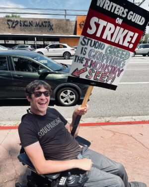 Zach Anner Instagram - As one of those writers and actors who works on several projects a year, but still can't make the minimum amount to get health insurance, this strike is pretty important to me! But it's not just about me obviously. It's about all the creative people who share their gifts and just want to make a living and be fairly compensated and appreciated for their work. #WGAStrong #sagaftra

Image description: Zach, sitting in his wheelchair on a sidewalk in Burbank. he wears a shirt that reads "SOULPANCAKE" and hold a sign. At the top, it says "writers guild of America on strike" and at the bottom, in hand-drawn, colorful letters, it reads "studio greed, is crippling the industry! See?!" And then has an arrow pointing towards Zach in his wheelchair.