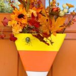 Meaghan B. Murphy Instagram - DIY candy corn 🌽 door hanger—don’t call it a wreath 🤣 HOW TO: ✂️ Cut a triangle out of foam ✂️ Cut yellow, orange & white strips out of craft foam paper. Tip: You can use tissue or construction paper if you’re hanging it indoors 🧵 Wrap the strips around the triangle and secure with sewing 🪡 pins 💐 Stick faux flowers in the top 🕷 Add a spider to make it truly spooktacular #wdcelebrates #wreathsofinstagram #wreathmaking #wreath #halloweendecor Halloween Headquarters