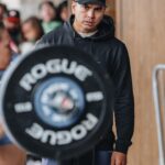 Tia-Clair Toomey Instagram - Sunday, 1:34pm: The fact that there is now a whole competition floor on the Dell Diamond concourse where a concession stand used to be — complete with 5 sets of double-under and lifting platforms, barricades and a sharp-looking @roguefitness backdrop — feels so natural, it’s easy to assume this was always the plan. It was not, but after four days of Book of Job-like weather, @caityhenniger and @billhenniger are taking no chances for the final event. Moving “The Cleanup” from the field to the concourse at the last minute is the latest example of the kind of logistical sorcery the @RogueInvitational has been performing behind the scenes all weekend. But not even Rogue can change the temperature, which dropped from 75° to 50° in the span of an hour earlier this morning. The top heat of women, the last of four heats, spend most of their 45 minute wait trying to stay warm; when @alexgazan_ 🇺🇸 takes a chair into the women’s bathroom, @laurahorvaht 🇭🇺 and @gabimiga 🇵🇱 follow suit. @tiaclair1 🇦🇺 remains outside, pacing around the warmup area with @shaneorr01’s jacket draped across her shoulders like a cape, superhero style. Shane, down to a just a longsleeve t-shirt, paces with her, shivering so hard he’s practically vibrating. A member of the Rogue event team notices, removes his own brand new Invitational hoodie and offers it to Shane, who accepts it with a mix of surprise and gratitude. By the time he pulls it on, @nick.p.jeffries is back at work, the only person on the concourse wearing shorts and a t-shirt. If you want to know what people mean when they say the Rogue Invitational goes above and beyond — it’s this. ✍🏽: @christinedca 📷: @emomphoto
