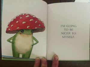Alison Pill Instagram - Oh my goodness it’s here!  @maybell.eequay has written the most wonderful book starring Little Frog!  It’s full of wisdom, and feels like a soul hug.  If you or someone you know is just having a tiiiime, this book will pick you up. (Image description: a blonde white woman in black tank on blue and white sofa holds a book called The Little Frog’s Guide to Self-Care)