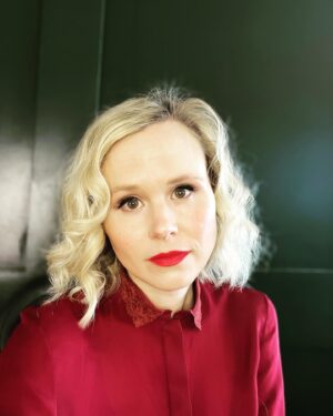 Alison Pill Instagram - Zoom press above and below. [image description 1: a blonde woman with curled hair wearing a red blouse and red lipstick. 2: a blonde Ewok- resembling dog lying on a cream blanket next to red chair legs and a pair of green Croc shoes]