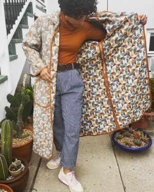 Jasika Nicole Instagram - I didn’t hop onto the quilted jacket trend til @byhandlondon released the #NerrisaCoat pattern a couple months ago. I loved the collar, the oversized fit and the long length, three elements that I had not previously seen in quilted coat patterns, and I had the *perfect* fabric for it- a geometric reversible quilted cotton smack in the middle of my color palette from @corefabrics. The pattern is very simple, pretty beginner friendly, and requires very little fit adjusting because it’s so oversized. I made the smallest size available (it goes up to a size E/ 61.75” bust) but it’s still incredibly roomy, like I could probably fit a sweater AND another coat under this lol. If I were to make it again I would probably slim down the fit by a couple inches and shorten it to shin length, which I might still do, but I wanted to get some wear out of this length before I chopped it up. I opted not to add buttons to the front and just have this be a kind of open jacket for layering. I made the bias binding myself and this is the only issue I had with the pattern- it says you need 5 meters of binding but I easily used 3 times that much for this view (I stopped counting after the third time I had to pull out my tools to make more tape 😵‍💫), so maybe that amount was meant for the shorter version of the jacket (because it doesn’t give different amounts for the different views). Thankfully I had just enough tencel scrap fabric to make all the tape I needed. The bias binding gives the jacket a nice pop of color while still feeling a bit neutral and I love how the colors in the fabric and tape play off of each other. I’m on the fence about the patch pockets on my version and think it would look even better with inset pockets, and because of the bulk of the quilted fabric, the drop sleeves look a little funky at the seams(I prefer inset sleeves). But those design choices are what makes this such a simple and quick pattern to finish, and I’m still happy with how this came out. The fabric is structured but soft and wearing it feels like you’re draped in a blanket cocoon- it actually makes a pretty great housecoat if you’re careful to not trip over it 😂