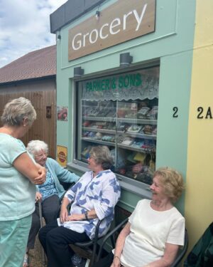 Anneka Rice Instagram - Oh my goodness. I went to Birkenhead, near Liverpool, today, to visit our Dementia Village, built on Challenge Anneka in 2022. It is flourishing. On a piece of wasteland a team of epic volunteers, builders, craftsmen, built an entire memory village - with pub, post office, record shop, cafe, bakers, cinema, bowling lawn, landscaped gardens! We worked in the pouring rain, in a recession, it was beyond stressful - but I thought my heart would burst, visiting it in the sunshine today, seeing what a difference it has made in this Wirral community. They now have over 400 volunteers helping out. The idea is that someone with dementia can wander safely round the village, dropping in on shops, listening to their favourite record, watching a film, having a singalong in the pub, doing whatever they want. It’s liberating for them and their loved ones because on every corner, sitting in the shops, offering them a cuppa in the cafe, there are carers to help them, chat to them, play bowls with them, sing with them. Each shop is curated to trigger memories. I went down a rabbit hole of memories seeing the Green Shield Stamps in the Post Office! IT IS BRILLIANT. I would love to see this rolled out across the UK. Well done @ageuk_wirral for having such vision 🩵🩵🩵 @age_uk #dementia @alzheimersresearchuk #village #ChallengeAnneka #charity #volunteers #community #joy @channel5_tv