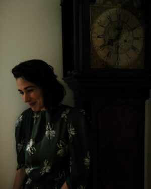 Geraldine Hakewill Instagram - outtakes w @rebeccahitchphotography @cathysavage 🕰️ 🪲 

ID: a woman with dark hair, wearing a green patterned dress, stands next to a grandfather clock. She is laughing.