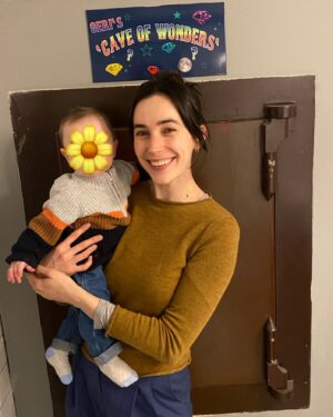 Geraldine Hakewill Instagram - Yes, that is a bank vault named “Geri’s Cave of Wonders”. Yes, it is named after me. Thank you, I will not be providing any context. 

ID: A woman with dark hair stands in front of a bank vault, holding a baby. She is smiling. Above the vault is a sign with multicoloured writing that says “Geri’s Cave of Wonders”.