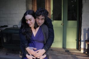 Geraldine Hakewill Instagram - It’s his first Father’s Day. I’m so happy my son has such a wonderful role model of how to be a compassionate, vulnerable, brave and loving man. And that he will always have such safe arms to be held in. My brother @lucasowen_ took this about a week before Robin arrived and we had no idea what was about to hit us. 

ID: A pregnant woman with long dark hair and a purple dress on sits, smiling, looking down. She is being hugged by a dark haired man who looks at the camera, his hands on her tummy.