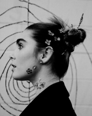 Maiah Stewardson Instagram - w love 🧚🏼‍♂️🌶☁️

[ID image 1: a black and white portrait of Maiah’s side profile. They have their hair in two space buns and delicate flowers lining their jawline and hair. 
Image 2: a portrait of Maiah wearing an all black coat with fluffy black slippers. They are standing with their feet close together and hands clasped 
Image 3: Maiah looks back at the camera. Their hands are lightly clasped and shadows hit the side of their body and face].
