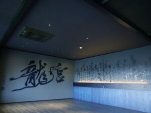 Mami Instagram - “龍宮”
2024,06,24, 佐賀県嬉野市・和楽園
@warakuen_chagokoro 

素晴らしい旅館の50周年を記念し、リニューアルされた客室の内装を担当いたしました。

「書く文字はお任せします」
と３代目よりオーダーをいただき、”日本三大美肌の湯” として知られる嬉野の泉質に癒されつつ、頭を振り絞って考えました。

左の壁「龍宮」は、
壁：艶消し藍色
インク：艶ありのネイビー
※これからベッドが来るので下のスペース空けてます💤

右の壁「亀ものがたり」は、
壁：艶消し藍色
インク：艶あり黒、艶ありネイビー、艶消し青緑

3色で書いた「亀ものがたり」は角度により見え方が変わり、まるで立体物のような平面作品を完成させることができました。

また、「龍宮」の文字を選んだ理由は以下。

--

①和楽園入り口にある池の亀たちが、竜宮城の乙姫を慕っていたという「亀ものがたり」のお話よりインスピレーションを受けた。

②竜宮城には水の神様がいる。和楽園はお茶と温泉を大切にしていて、どちらも水が鍵となる。

③竜宮城に赴いた者へは宝物が与えられる。私自身も、和楽園にて沢山の宝物となる時間を過ごさせていただいた。

④和楽園から徒歩圏内に豊玉姫（別名：乙姫）をお祀りする神社があり、書く前に参拝した。

〜三代目より個人的なエピソードも入れてOKと言われたので〜

⑤和楽園に伺った２日間、爽やかな雨が降っていて、龍神様が喜んでいるように感じた。

⑥自分自身の中にも新しい命がいて、水（羊水）がとても大切。龍年に子供を授かり出産することと、和楽園も今年50周年を期に新たな挑戦をするということで、「竜宮」を「龍宮」と表記した。

--

和楽園さん50周年おめでとうございます。
そしてお招きいただき、特別な時間を作っていただき、ありがとうございます！！！
またすぐに行きたい♨️🍵　

#和楽園 #嬉野温泉 #嬉野茶
#書道家万美 #mamimozi #Calligraf2ity