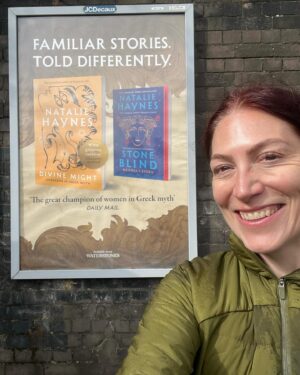 Natalie Haynes Instagram - It is such a thrill to see these posters out in the wild! Thank you to @picadorbooks for this amazing campaign 🧡🧡 Please note lack of glasses because on my way back from the gym. Bonus fact: I asked a stranger to take this pic in the 1 minute before her train arrived, and she said yes