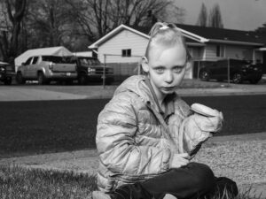 Robin de Puy Instagram - Emily (6), Twin Falls, Idaho 💕
 
Drowned in dark thoughts I wander through a big Walmart in Twin Falls, Idaho. It is the intense gaze of a young girl that brings me back to earth. In times of despair, the look of the kid is comforting as well as painful: the ultimate imagination of innocence, which only a child can show. Sitting in a shopping cart, being pushed by her big sister, the world is passing by.
 
I would love to photograph the girl. I nervously approach them, scared of being rejected. “Is your mom here too?”, I ask. “She's in the restroom, but she will be back soon”, the sister says. Meanwhile, the bright blue eyes of the little girl are staring right through me. I try to keep the conversation going, without appearing to be a creepy stalker. When the mother joins us, the big sister has already found me on Instagram and wants to take a selfie with me. Mom is keeping an appropriate distance and I see her – understandable – concerns. I give her my phone number and await. Not much later I receive the message I was hoping for: “You can come over.”
 
The name of the girl is Emily. Her intellectual disability sometimes makes things harder, but because of the loving presence of big sister Kenzie (15), brother Lucas (8), mom Ashley (40) and dog Piper she can move quite carelessly through life. In contradiction with a lot of struggling parents, her mom has, with the help of school and doctors, paved the way through the complex American bureaucracy with as a result a happy, healthy kid in school. “Without the help, I could not have done it.”
 
Like a little kitten, Emily plays in the grass. Continuously she is approaching me closer and closer, followed by running away or hiding in her pink jacket. Everything is sweet about her. Her light, fine hair, the breadcrumbs on her chin, the small hands which are holding a (still frozen) peanut butter and jelly sandwich, the nearly rolling tear that arises because of the wind and the accompanying booger.
An innocent child. Freely she rolls through the grass, and I roll with her. I want nothing more than to forget or not know – it works.