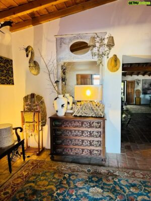 Rui Oliveira Instagram - Hoje apeteceu-me redecorar o hall do monte. Aqui o tempo é mais tempo, dá para tudo. Gosto do jarrão Bordalo Pinheiro a que juntei uns galhos de oliveira para que uma andorinha neles pudesse “pousar”. Não sendo aficionado  deslumbro-me, contudo,  com a opulência e minúcia dos bordados dos trajes de toureio. A casaca foi-me ofertada pelo cavaleiro Rui Fernandes, no “Você na Tv”, já o “traje de luces” pertenceu ao jovem toureiro monfortense João Silva “El Juanito” e foi-me dado no programa da tarde. Ambas as peças evocam memórias de colhidas pelo que se tornaram malditas para os dois protagonistas da festa brava. A cómoda,  em segunda mão,  foi um achado numa venda da Emaús, pelo trabalho da madeira da sua frente. A rusticidade das peças: do espelho de aspecto provençal ao candeeiro feito de troncos, passando pelos cajados que jazem no pote cerâmico, usados na condução do gado e para apoio do pastor, pelo antigo cesto de piquenicar onde agora se guardam alguns bonés e agasalhos de pescoço, para serventia sempre que necessária, e pelas cabeças em madeira que aludem a uma prática comum na região , dão o mote para a restante decoração de todo o monte. É Alentejo. E depois eu gosto de decoração.