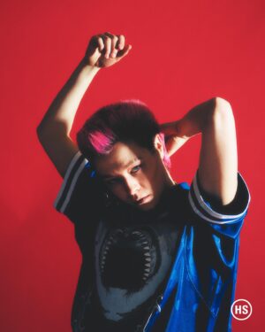 Emma D'Arcy Instagram - a cross boy gets his picture taken for @highsnobiety !! 🗯️

masc blessings by @rosefordestudio 
photographed by @petrosstudio 
crown by @jodytaylorhair 
mug by @justinejenkins 
pink by @elenitses @nudostudio.ldn 
production by @_kill_hill 
  wonderful chat from @willahbennett 

love and power, always @dobbscld and @dfm16

[image description: a gallery of two photographs. The first image shows Emma, a white, androgynous non-binary person, with spiked pink hair, photographed against a red background. They look to camera. Half of their face is in shadow. Emma wears an electric blue football shirt, with white piping around the collar. They have a thick twisted silver chain around their neck. From their left ear, a silver hoop hangs in silhouette against the red background. In the second image, Emma wears the same outfit. This time, a shark’s open jaws are visible on the chest of the blue shirt. Emma has their arms above their head. One hand brushes the back of their neck. The other hangs in the air, fingers caught in the light.]