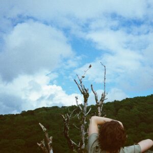 Emma D'Arcy Instagram - ☁️ @tmayb 

[image description: The camera looks out over dense woodland that meets a blue, cloud-patterned sky. In the foreground are the bare, greyish branches of a tree, and the torso of a person leaning against the tree, looking toward the sky.]