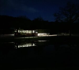 Emma D'Arcy Instagram - [image description: at night, the camera looks out over a lake towards a line of trees that are silhouetted against a dark, cobalt blue sky. In front of them, an empty, stationary bus is illuminated from the inside, casting a pool of greenish light around it. The bus is reflected upside-down in the water.]