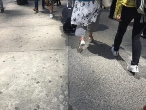 Emma D'Arcy Instagram - [image description: a gallery of two photographs, taken in New York. The first image shows the legs of pedestrians walking on a brightly lit sidewalk. The camera points down towards the illuminated concrete pavement, which is patterned with dark spots of chewing gum. The legs of the pedestrians are in motion; a suitcase is pulled away from camera, a light cotton shirt catches the breeze. The second image show three pairs of legs walking on a brightly lit sidewalk. Two pairs of legs in the centre of the frame are walking towards the camera. The bodies of the pedestrians cast graphic shadows on the concrete. A hand holds an iphone. The road, to the left of frame, reads ‘BUS’.]