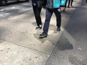 Emma D'Arcy Instagram - [image description: a gallery of two photographs, taken in New York. The first image shows the legs of pedestrians walking on a brightly lit sidewalk. The camera points down towards the illuminated concrete pavement, which is patterned with dark spots of chewing gum. The legs of the pedestrians are in motion; a suitcase is pulled away from camera, a light cotton shirt catches the breeze. The second image show three pairs of legs walking on a brightly lit sidewalk. Two pairs of legs in the centre of the frame are walking towards the camera. The bodies of the pedestrians cast graphic shadows on the concrete. A hand holds an iphone. The road, to the left of frame, reads ‘BUS’.]