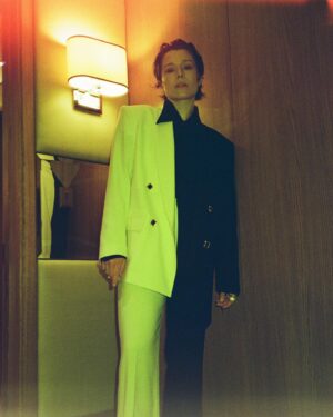 Emma D'Arcy Instagram - 🔥 Happy bloomsday !

🔥 Photos by @tmayb 

Two-tone suit by @canaku_ with @bitter.000000 grills and @boucheron diamonds 
Brought together by genius @rosefordestudio 
Hair always by @jodytaylorhair 
and makeup by brilliant @phoebewalters 

#hotd 

[image description: a slim non-binary person is standing against a wooden door by a cream, wall-mounted lamp. They wear a double-breasted suit that is half black and half white. They wear a black shirt, the pointed collar of which sticks out over the white lapel of the blazer.]