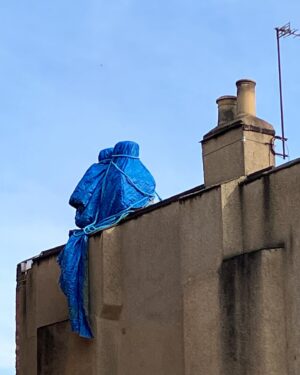 Emma D'Arcy Instagram - [image description: The camera is looking at the very top of a beige building, against a dusty blue sky. There are two sets of chimney stacks. The chimney stack on the right is rendered in the same brownish beige as the rest of the building. The chimney stack on the left, has been covered with an electric blue tarpaulin. A blue rope binds the tarpaulin to the chimney, creating the shape of two bodies, nestled close together. The end of the blue tarpaulin hangs off the edge of the roof, like a blanket hanging off a bed, or the tail of a cat.]