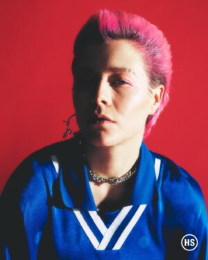 Emma D'Arcy Instagram - a cross boy gets his picture taken for @highsnobiety !! 🗯️

masc blessings by @rosefordestudio 
photographed by @petrosstudio 
crown by @jodytaylorhair 
mug by @justinejenkins 
pink by @elenitses @nudostudio.ldn 
production by @_kill_hill 
  wonderful chat from @willahbennett 

love and power, always @dobbscld and @dfm16

[image description: a gallery of two photographs. The first image shows Emma, a white, androgynous non-binary person, with spiked pink hair, photographed against a red background. They look to camera. Half of their face is in shadow. Emma wears an electric blue football shirt, with white piping around the collar. They have a thick twisted silver chain around their neck. From their left ear, a silver hoop hangs in silhouette against the red background. In the second image, Emma wears the same outfit. This time, a shark’s open jaws are visible on the chest of the blue shirt. Emma has their arms above their head. One hand brushes the back of their neck. The other hangs in the air, fingers caught in the light.]