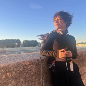 Rosanne Mulholland Instagram - Ela faz cara de plena, mas tá agarrada no vinho. 🍷

Bisous.

#PraTodosVerem Rosanne, uma mulher branca de cabelos castanhos curtos, está encostada em uma mureta ao pôr do sol segurando uma garrafa de vinho tinto.