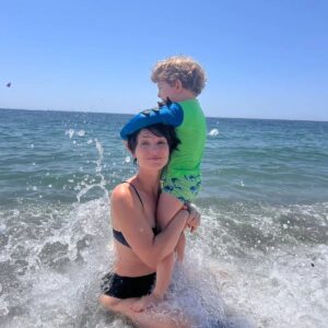 Rosanne Mulholland Instagram - Ah, a maternidade e as férias....
Bisous.
💛

#PraTodosVerem Uma sequência de três fotos na praia. Na primeira, o Davi, menino branco  de 3 anos com roupa de praia verde, está em pé cima das pernas da mãe, Rosanne, mulher branca de biquíni azul marinho, que está sentada na parte rasa da praia, onde a água cobre as pernas. Davi está se pendurando em sua cabeça enquanto ela mantém um sorriso meio desesperado. A segunda é uma variação da primeira. O sorriso não mostra os dentes e é mais debochado. Uma onda bate nas costas de Rosanne. Na terceira foto,  a água recuou e vemos as pedrinhas da praia. Davi está pendurado de forma que seu rosto está escondido atrás da cabeça da mãe. Rosanne agora dá um sorriso sincero. Fim da descrição.