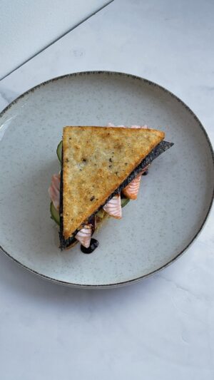 Isadee Jansen Instagram - SUSHI SANDWICH 

Ingrediënten voor 1 sandwich:
- 100 g sushi rijst
- 1 tl rijstazijn
- 0,5 tl suiker
- Snufje zout
- 1 tl furikake

Voor de tonijnsalade:
- 50 g tonijn
- Kewpie mayo
- Sojasaus
- Sesamolie
- Wasabi
- Sesamzaadjes
- Bosui

Overige ingrediënten:
- 4 plakjes zalm sashimi
- Komkommer
- Avocado
- Unagi saus
- Nori vellen
- Neutrale olie
- Vershoudfolie 
- Gasbrander

Bereidingswijze:
1. Kook de sushi rijst volgens de instructies op de verpakking.
2. Voeg aan de warme rijst de rijstazijn, suiker, een snufje zout en furikake toe. Laat de rijst afkoelen.
3. Bedek een bakplaat of ovenschaal met vershoudfolie en verdeel de rijst erop. Druk het plat tot een laag van ongeveer 1 cm dik. Verpak de rijst in folie en plaats het in de vriezer. LET OP: druk de rijst goed aan, anders kan het uit elkaar vallen tijdens het frituren. En je wilt liever dat de rijst wat natter dan droger is, zodat het goed aan elkaar blijft plakken. 
4. Snijd de tonijn in kleine stukjes en meng met kewpie mayo, sojasaus, sesamolie, wasabi, bosui en sesamzaadjes.
5. Snijd de komkommer en avocado in plakjes. Knip de nori vellen in driehoeken. Haal de bevroren rijst uit de vriezer en snijd in driehoeken.
6. Verhit neutrale olie en frituur de rijst tot deze goudbruin en krokant is. Laat uitlekken op keukenpapier. 
7. Besmeer de gefrituurde rijst toast met kewpie mayo. Voeg vervolgens een nori vel, de tonijnsalade, komkommer, avocado en de zalm sashimi toe.
8. Gebruik een gasbrander om de zalm te flamberen. Bestrijk met unagi saus, leg er een tweede nori velletje op en sluit af met de tweede rijst toast.

Selamat makan!